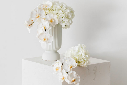 Custom made artificial floral arrangement featuring white hydrangeas and orchids in a white textured ceramic vase.