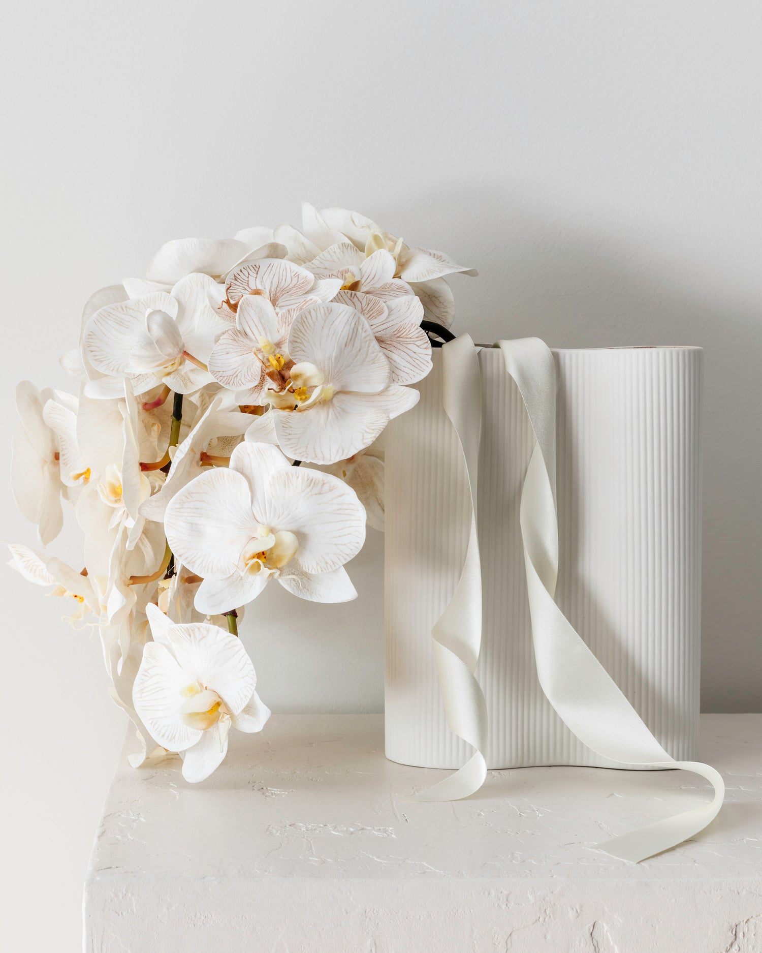 Stunning white real-touch orchids arranged in a textured ceramic white vase, elegantly displayed on a plinth.