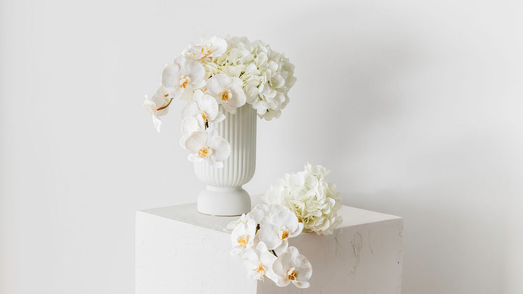 Custom floral arrangement featuring orchids and hydrangeas in a white, textured ceramic vase, displayed on a plinth.